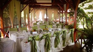 Un área de comedor al aire libre con mesas dispuestas para una comida, cubiertas con manteles blancos y fajos verdes, bajo una pérgola de madera con cortinas.