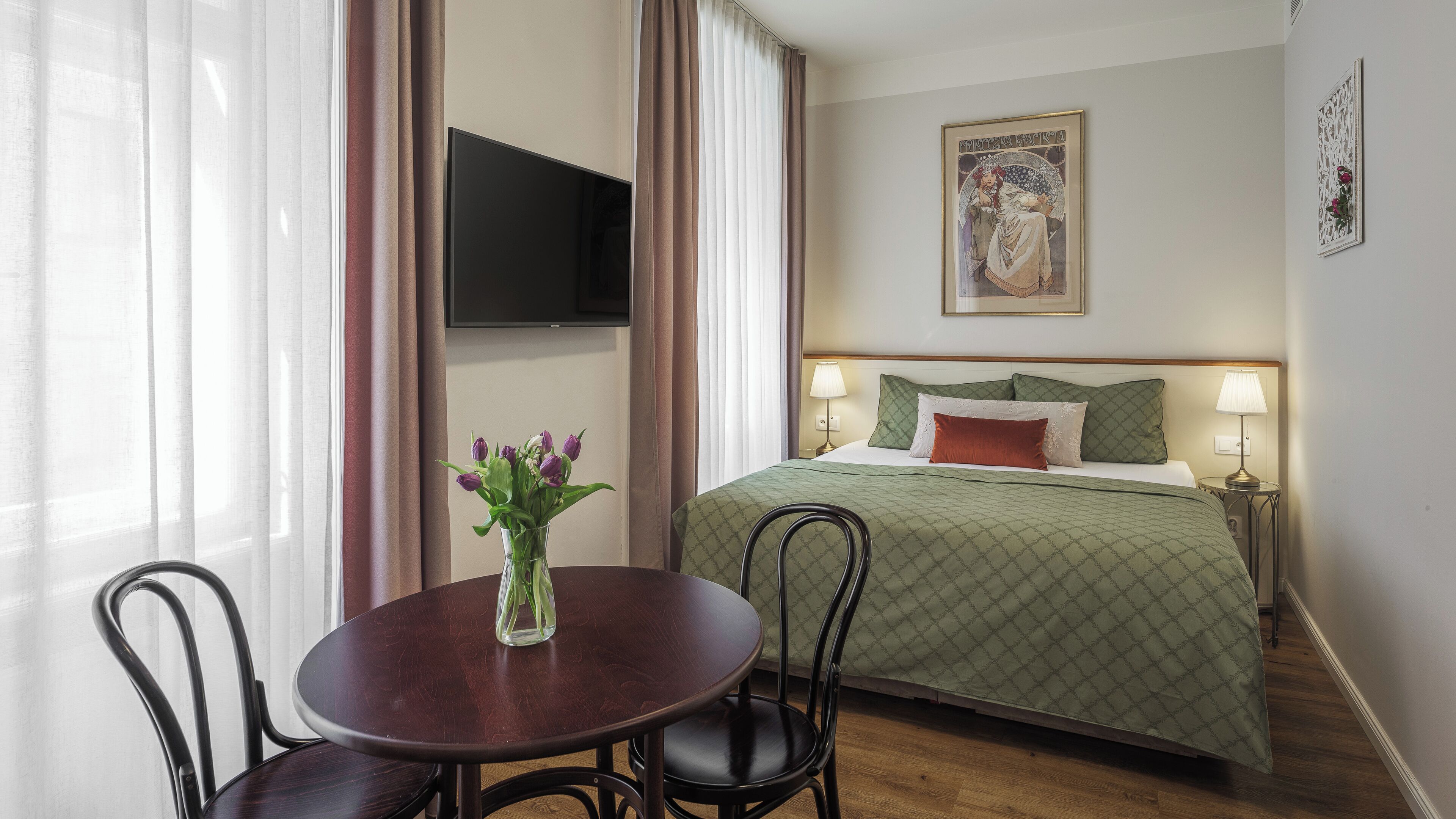 A hotel room with a bed, a TV mounted on the wall, a table with chairs, and a vase with flowers.