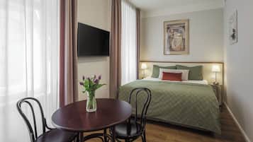 A hotel room with a bed, a TV mounted on the wall, a table with chairs, and a vase with flowers.