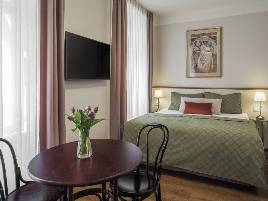 A hotel room with a bed, a TV mounted on the wall, a table with chairs, and a vase with flowers.