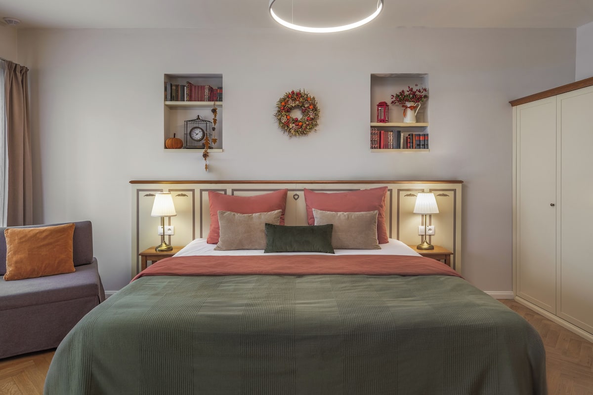 A bedroom with a bed, bedside lamps, a nightstand, a chair, a bookshelf, a decorative wreath, and a wardrobe.