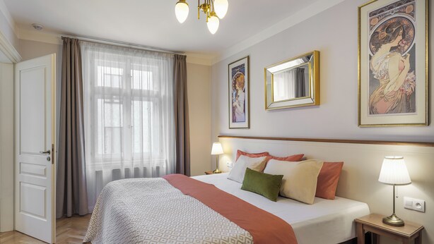 Una habitación de hotel con una cama, una mesita de noche con lámpara, una ventana con cortinas y obras de arte en las paredes.