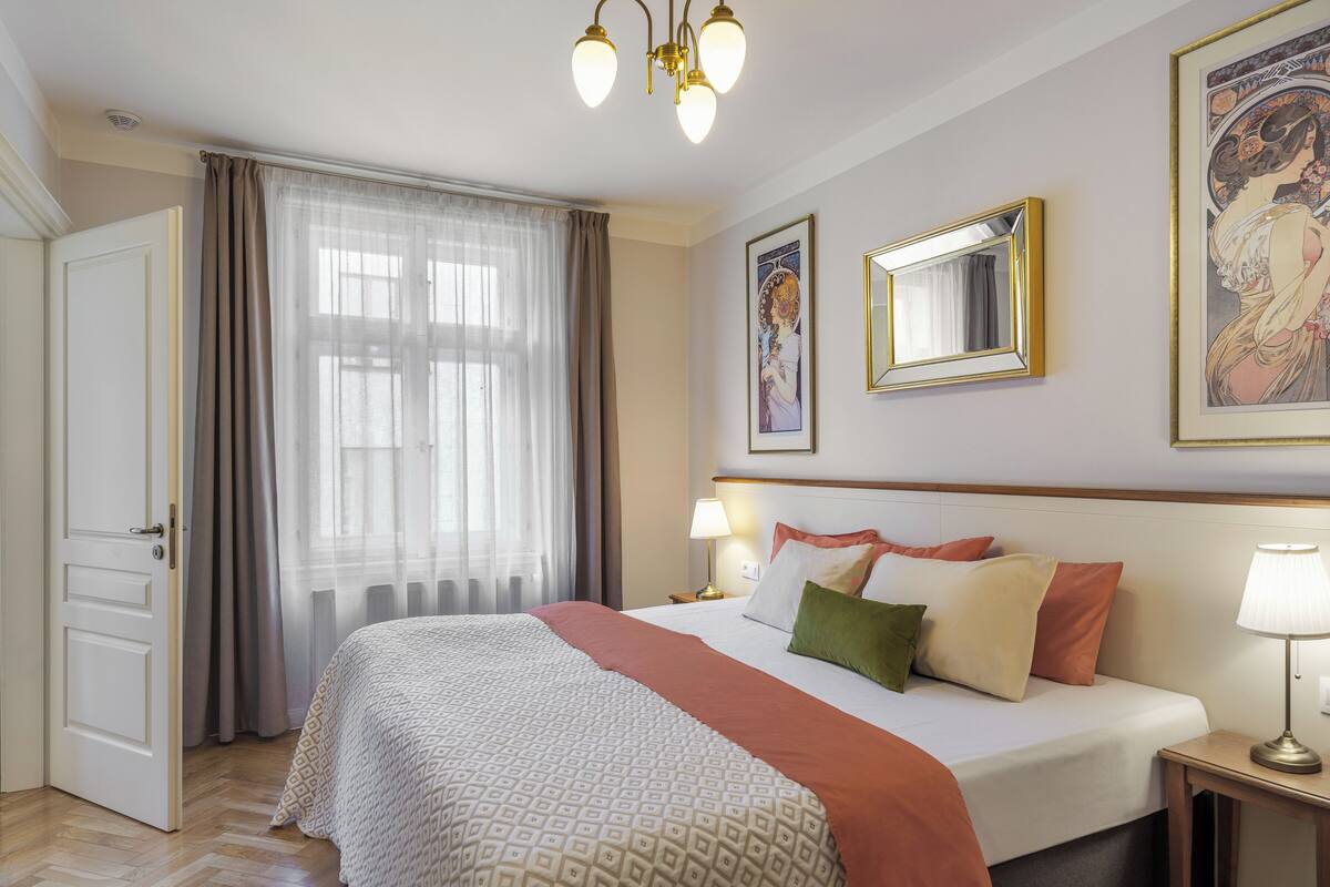 A hotel room with a bed, a bedside table with a lamp, a window with curtains, and artwork on the walls.