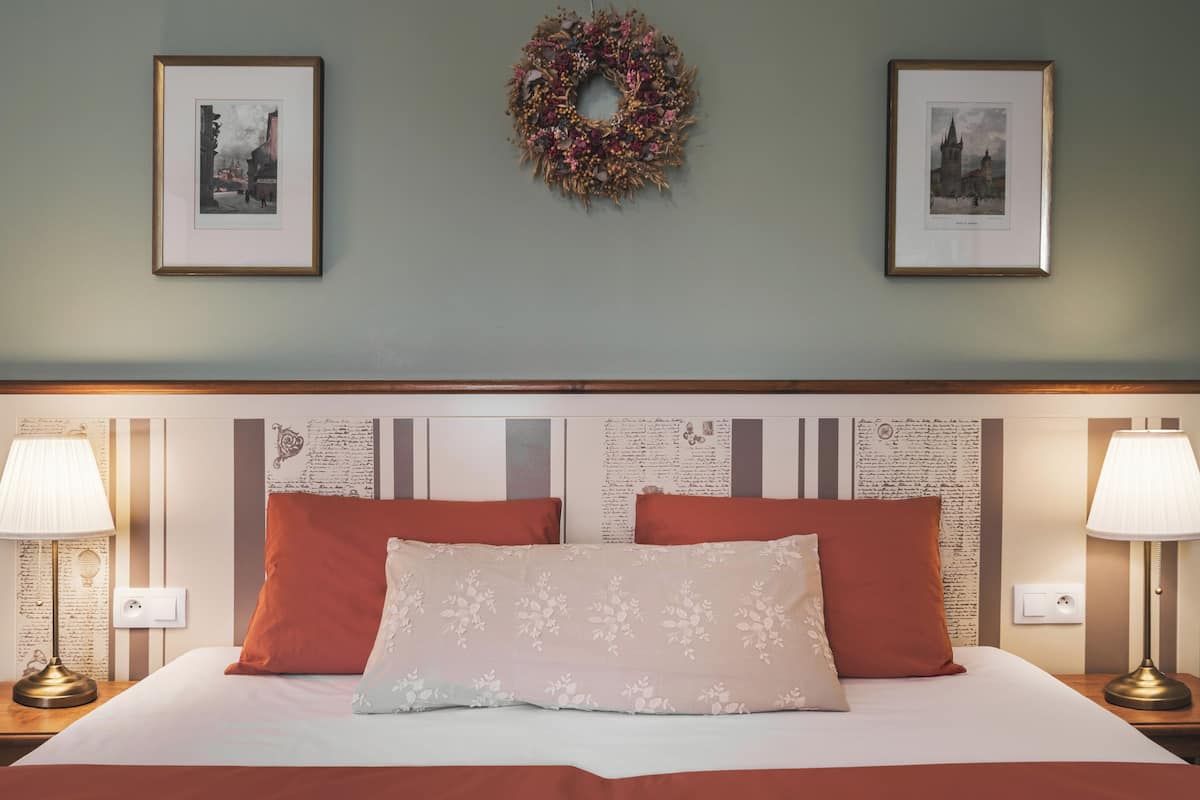 A neatly made bed with a floral pillow, two red pillows, and a white bedspread. There are two bedside lamps on either side of the bed, and two framed pictures above the bed.