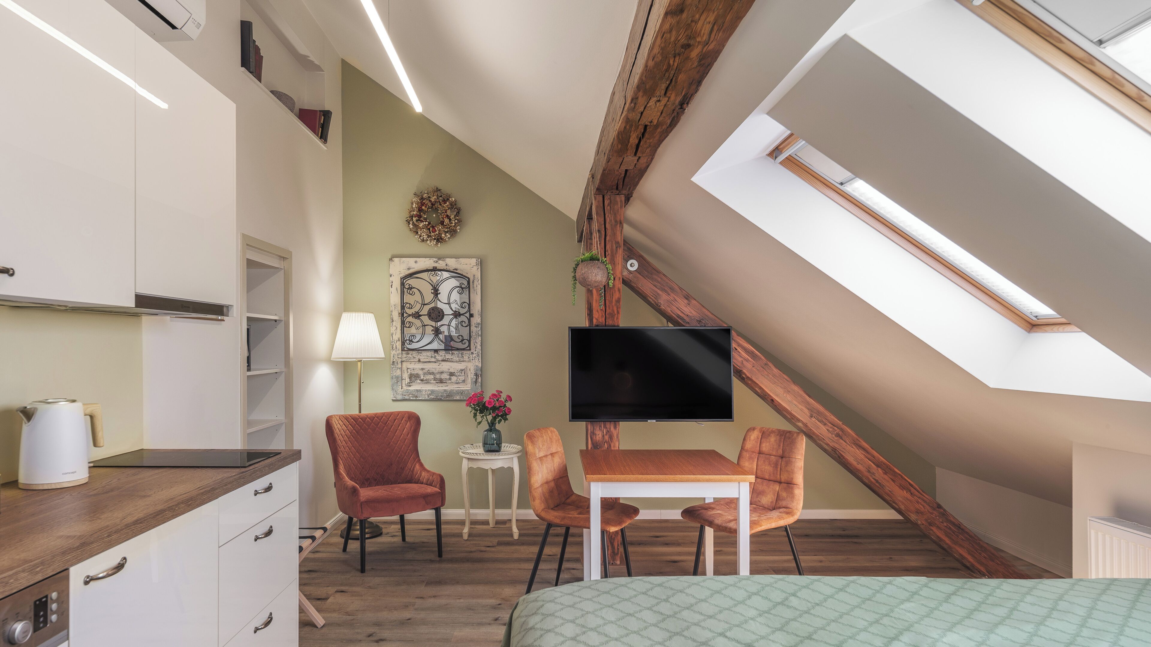 A compact room with a kitchenette, a flat-screen TV, a dining table with chairs, a bed, and a skylight.