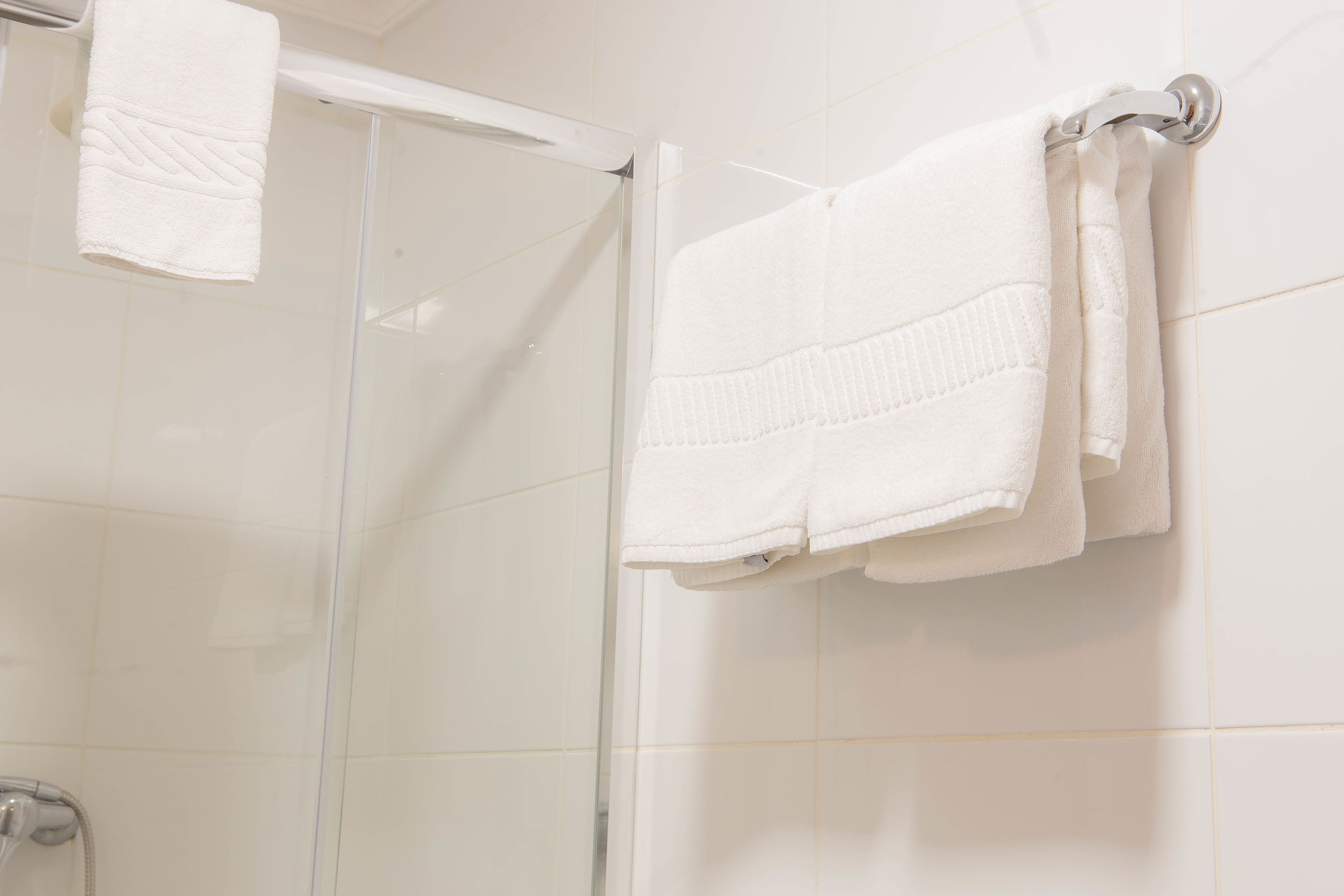 A bathroom with white tiled walls, a glass shower enclosure, and two white towels hanging on a metal towel rack.