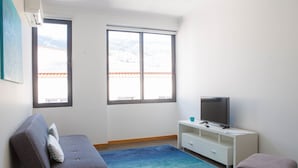 A room with a sofa, a TV on a white stand, a window with a view of mountains, and a blue carpet. - Five Design Rooftop by Storytellers (Funchal)