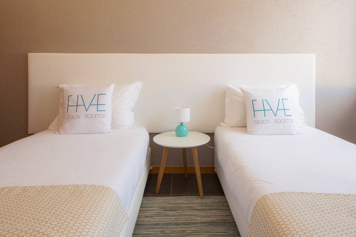 A hotel room with two single beds, each with a pillow that reads "HAVE DESIGN ROOFTOP". There is a small round table with a lamp between the beds.