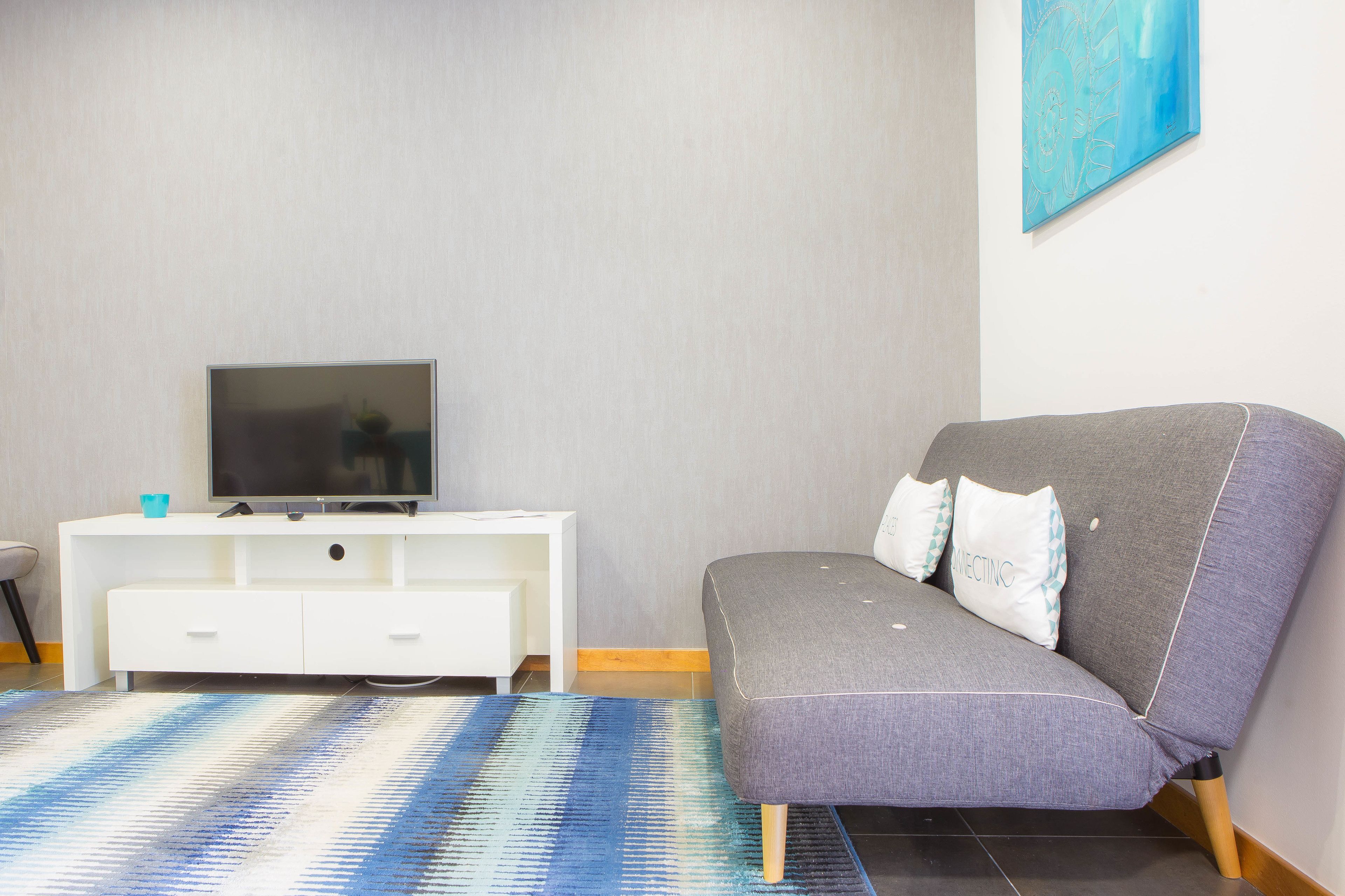 A modern living room with a grey sofa, a flat-screen TV on a white cabinet, and a patterned rug.