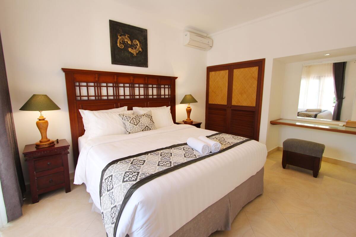 A neatly made bed with a patterned bedspread and pillows, two bedside tables with lamps, a small bench, and a mirror above the bench.