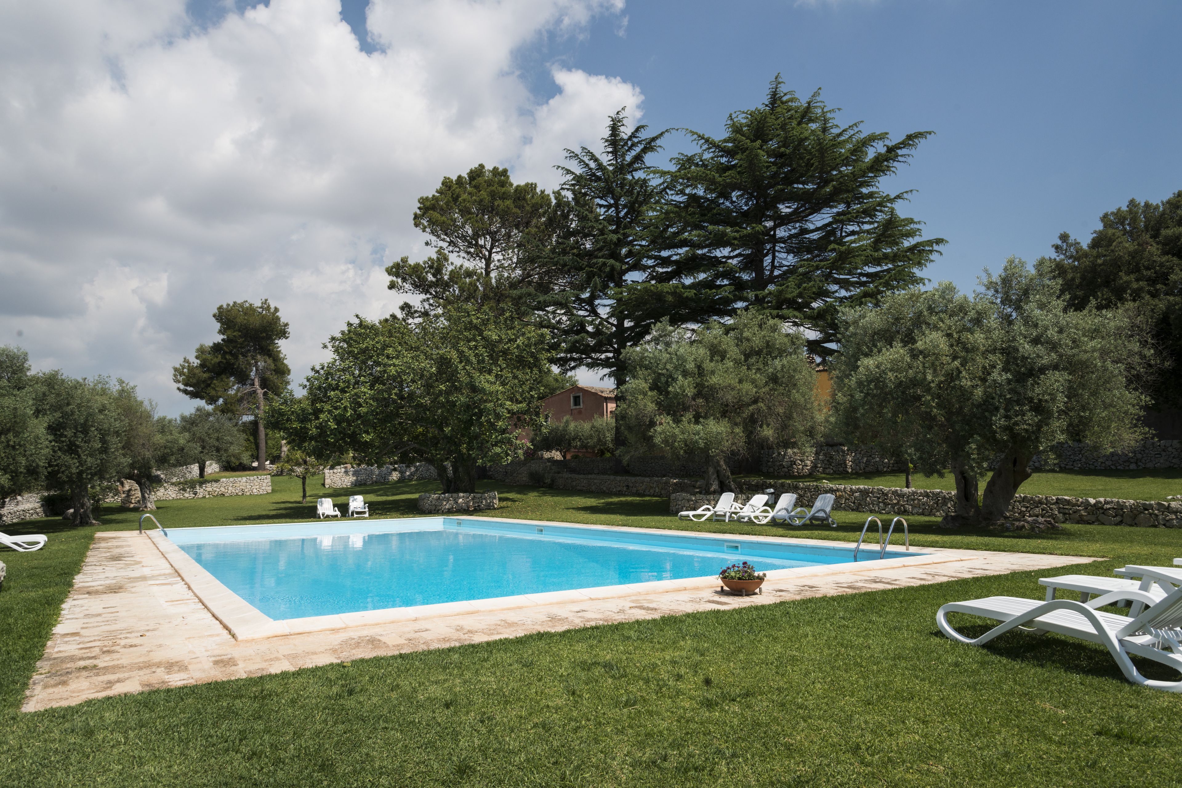Piscina rectangular con borde de piedra, rodeada por un césped cuidado y varias sillas de descanso blancas.