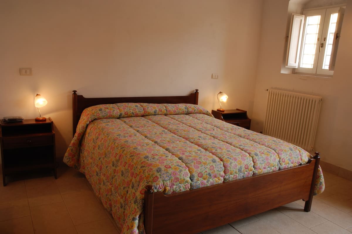 Une chambre à coucher avec un lit en bois, deux tables de chevet, une commode, un radiateur et une fenêtre donnant sur un beau paysage.