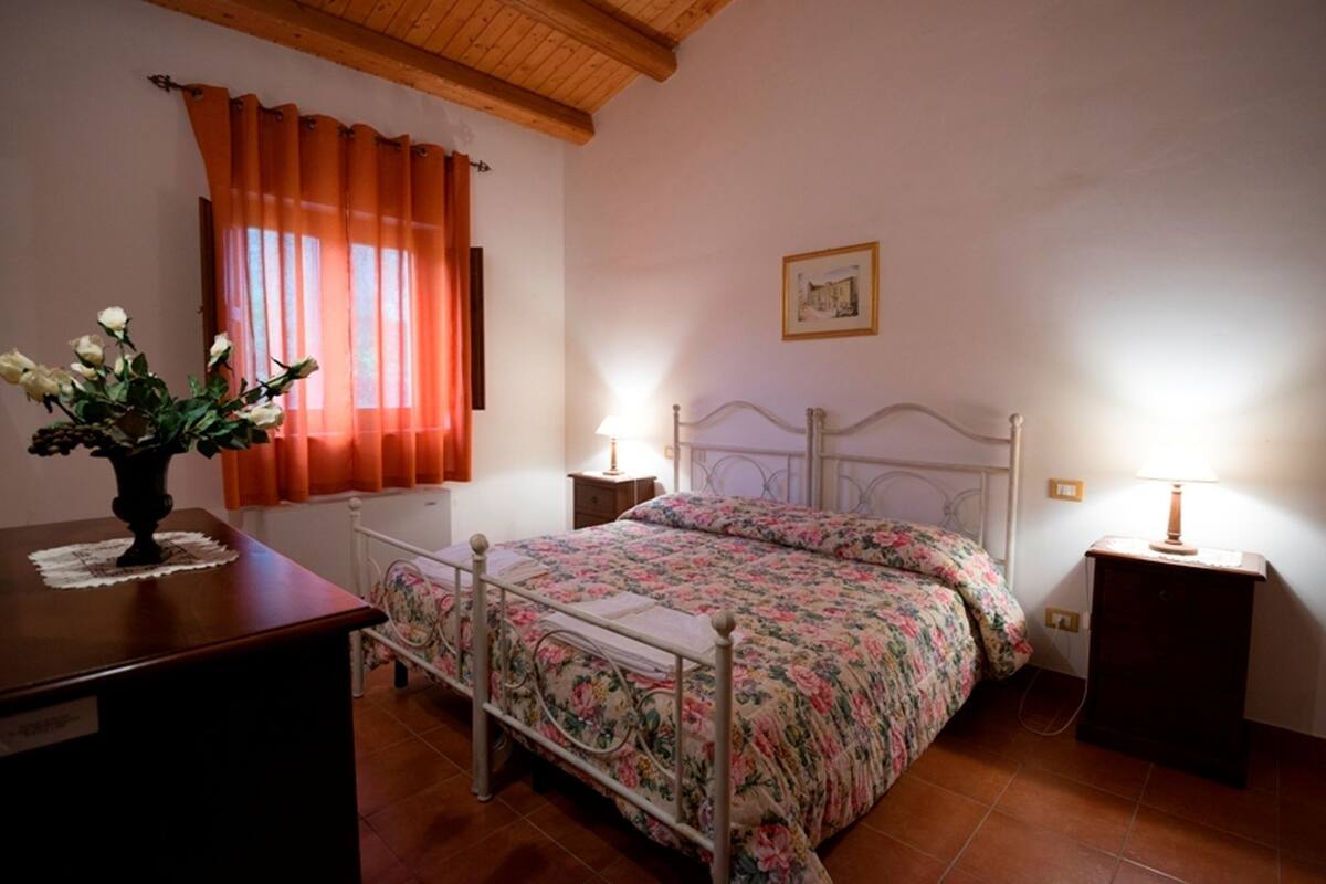 Une chambre à coucher avec un lit, deux tables de chevet, une petite table, une commode en bois, un vase avec des fleurs et une fenêtre avec des rideaux orange.