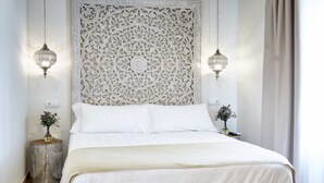 Una cama con cabecera blanca, dos lámparas de pared y una mesita de noche con un florero.