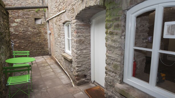 A narrow stone corridor with a small table and chairs, a door, and a window with a view into a room. - The Priory Cellars Abbots Leigh (Bristol)