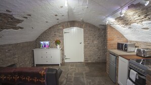 A compact kitchen with a stone wall, a white door, a microwave, a stove, and a small refrigerator. - The Priory Cellars Abbots Leigh (Bristol)