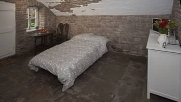 A bedroom with a stone wall, a bed with a patterned bedspread, a small table with fruit, a chair, and a window with a view.