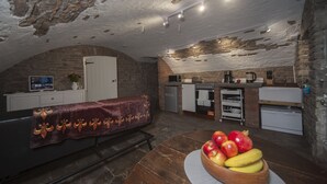 A cozy, rustic kitchen with stone walls, a wooden dining table with fruit, and a massage table with a blanket. - The Priory Cellars Abbots Leigh (Bristol)