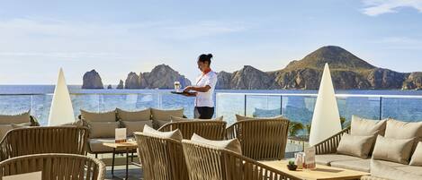 A terrace with wicker furniture, a waiter serving, and a view of the sea and rocky landscape.