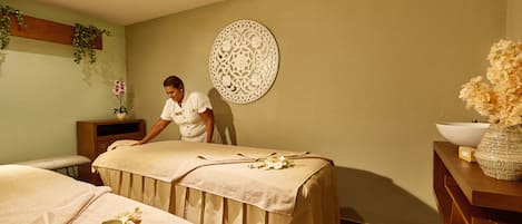 Un cuarto de spa con dos camillas de masaje, un adorno en la pared y un mueble de madera con un jarrón de flores.