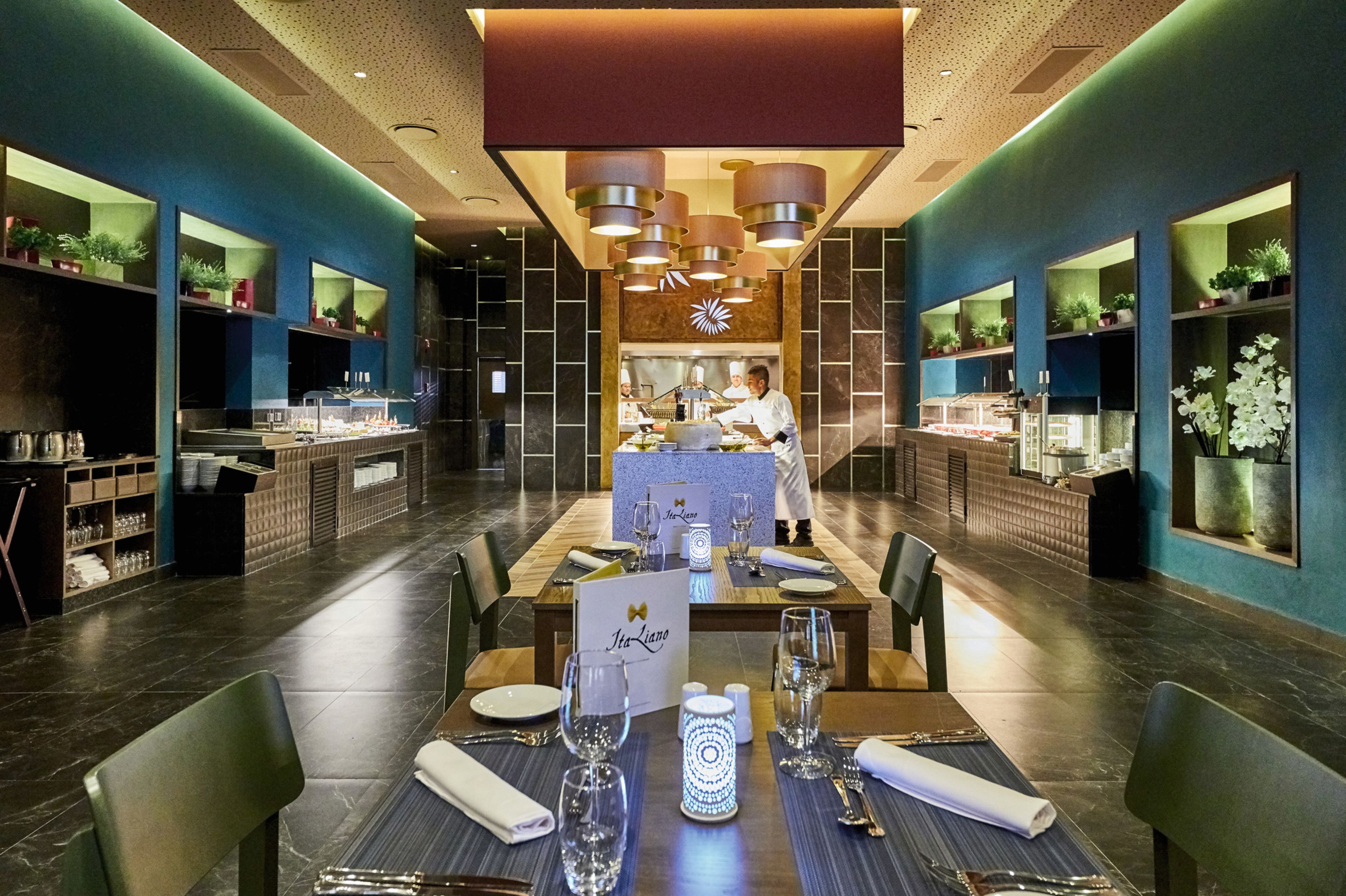 Ein Restaurant mit modernem Interieur, das einen zentralen Essbereich mit gedeckten Tischen, eine Küche im Hintergrund und dekorative Pflanzen zeigt.