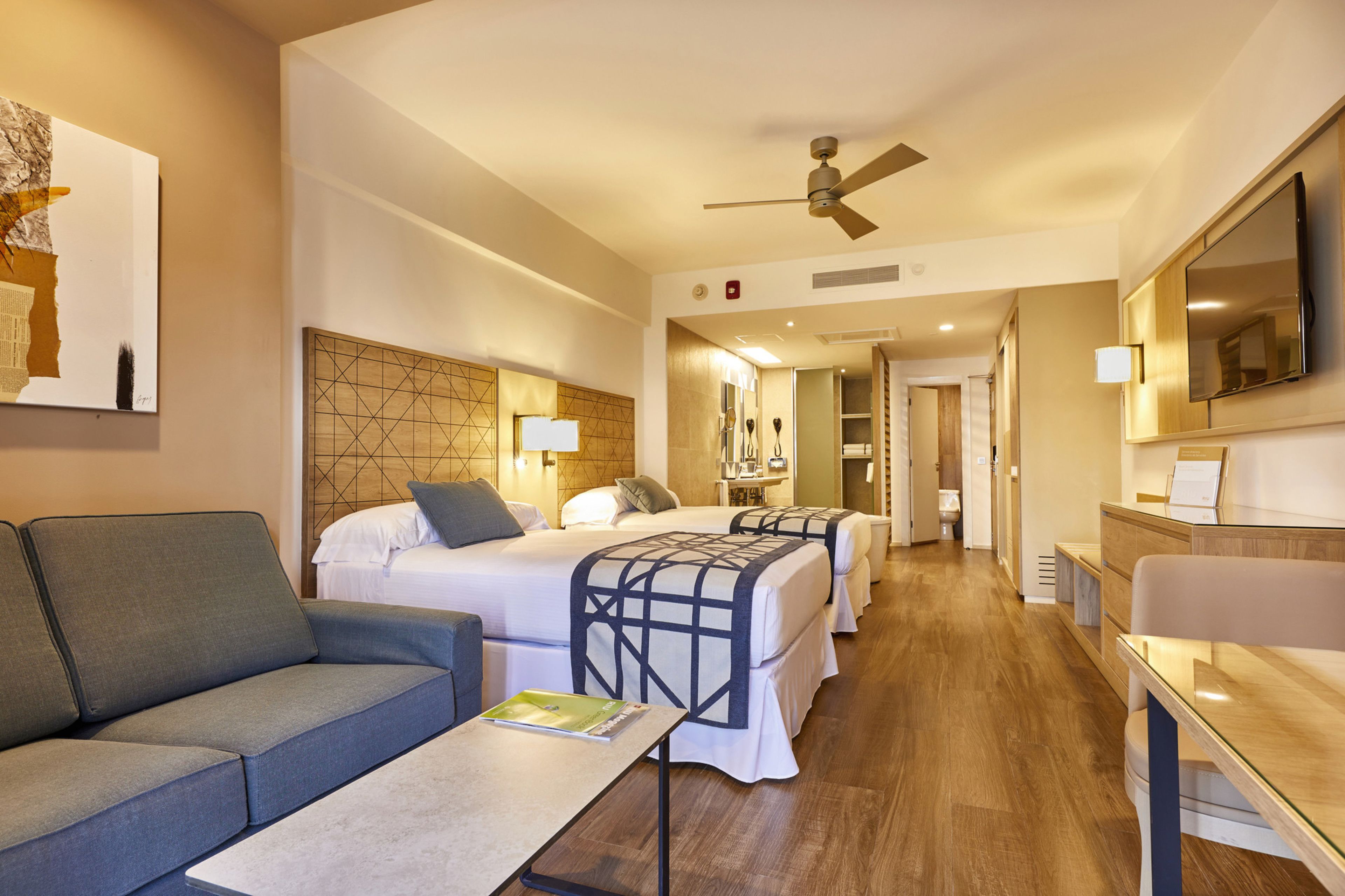 Une chambre d’hôtel avec un canapé, deux lits avec des têtes de lit, une télévision, un ventilateur de plafond et un plancher en bois.