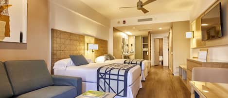 Une chambre d’hôtel avec un canapé, deux lits avec des têtes de lit, une télévision, un ventilateur de plafond et un plancher en bois.