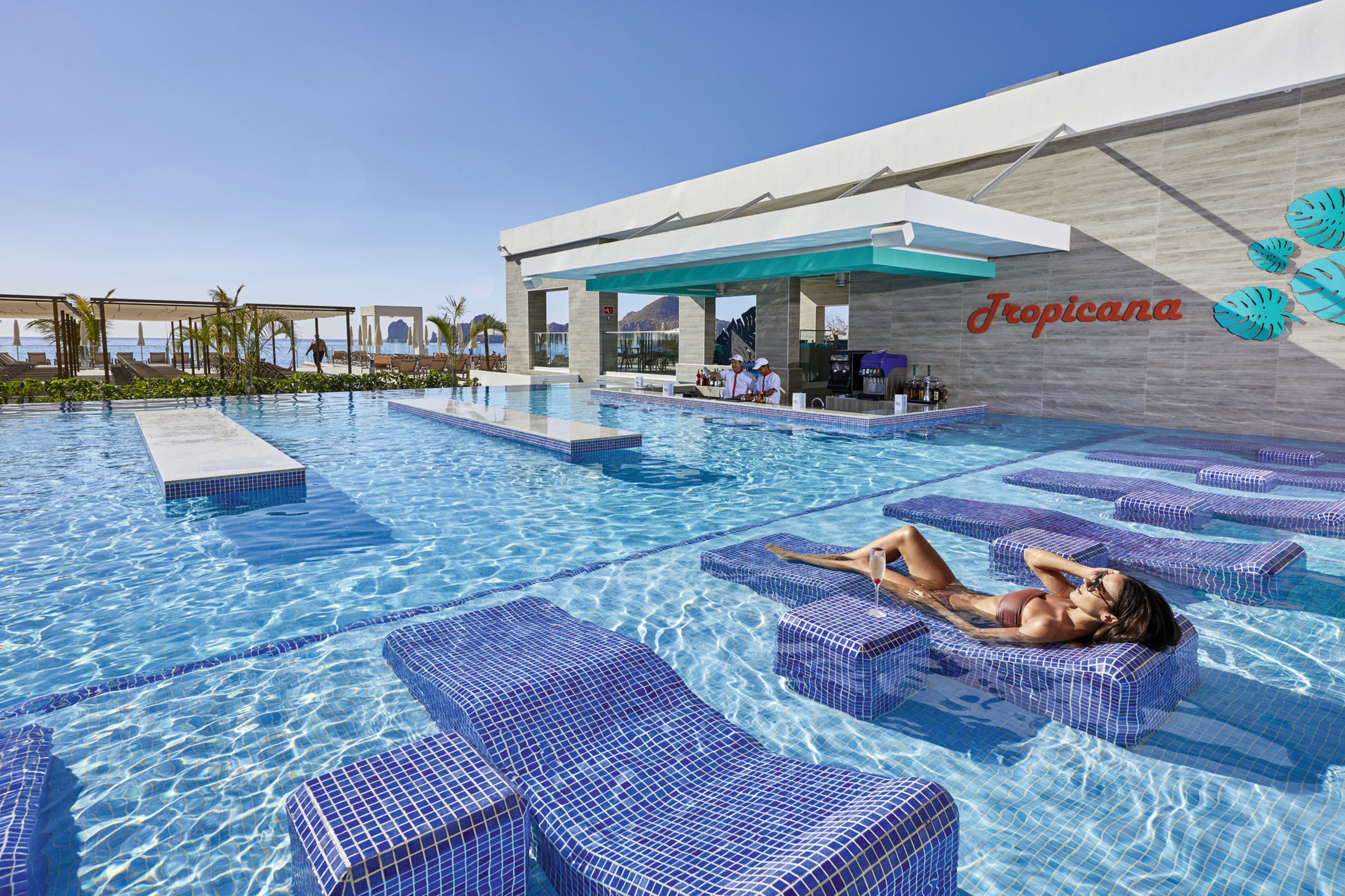 Zona de piscina con sillones de descanso y un bar, con el nombre 'Tropicana' a la vista en la pared.