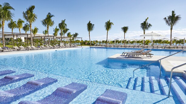 A swimming pool with clear blue water, lounge chairs, and palm trees. - Hotel Riu Palace Costa Mujeres - All Inclusive (Costa Mujeres)