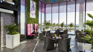 A modern lounge area with wicker furniture, a large window with a view of the beach, and a green wall with a floral painting. - Hotel Riu Palace Costa Mujeres - All Inclusive (Costa Mujeres)