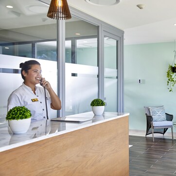 A reception area with a receptionist, a wooden reception desk, potted plants, wicker chairs, and hanging plants on the wall.