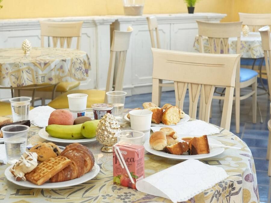 Mesa de jantar posta com diversos alimentos e bebidas, rodeada por cadeiras e uma toalha de mesa estampada.