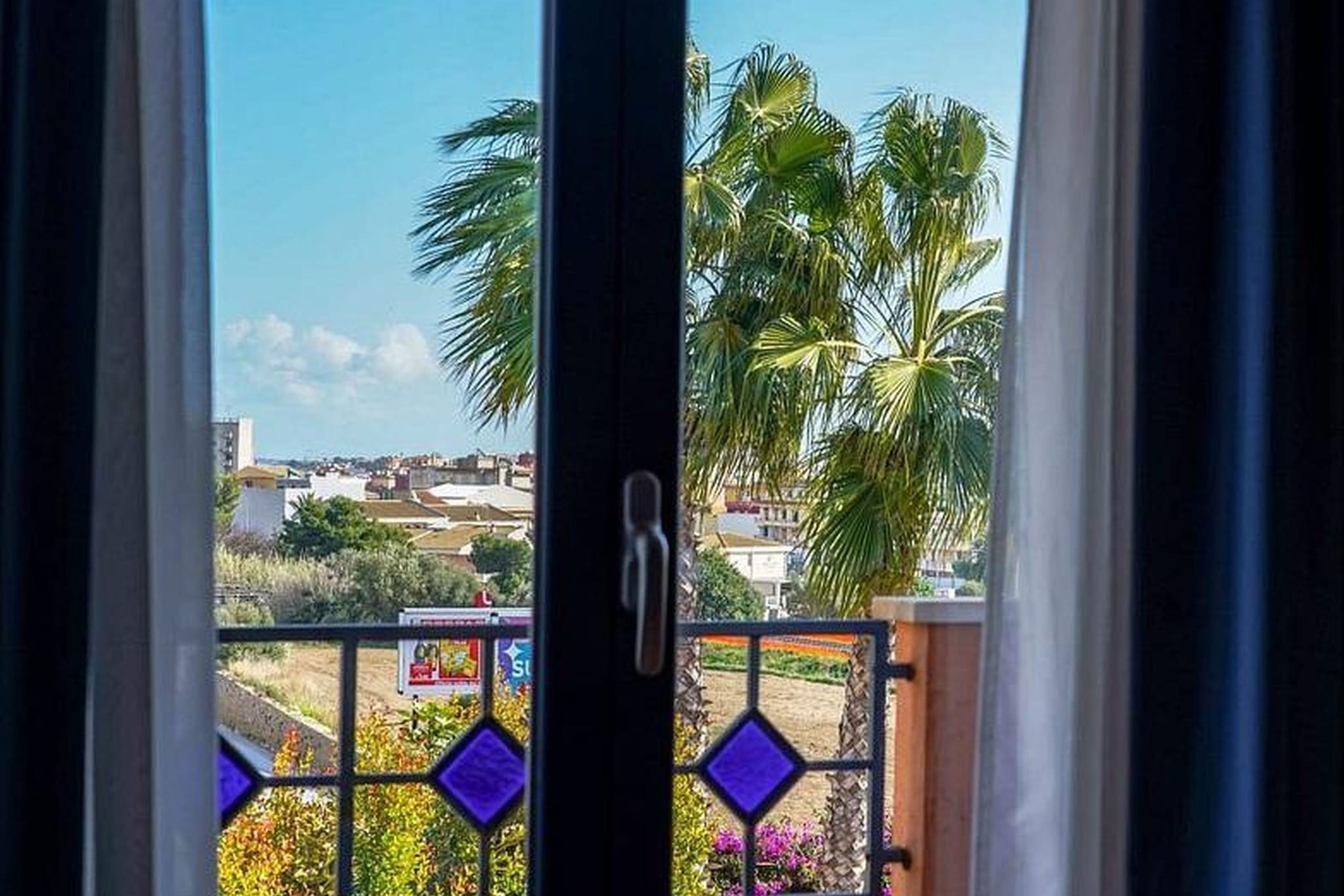 A view through a window with a metal gate, featuring a clear sky and palm trees.