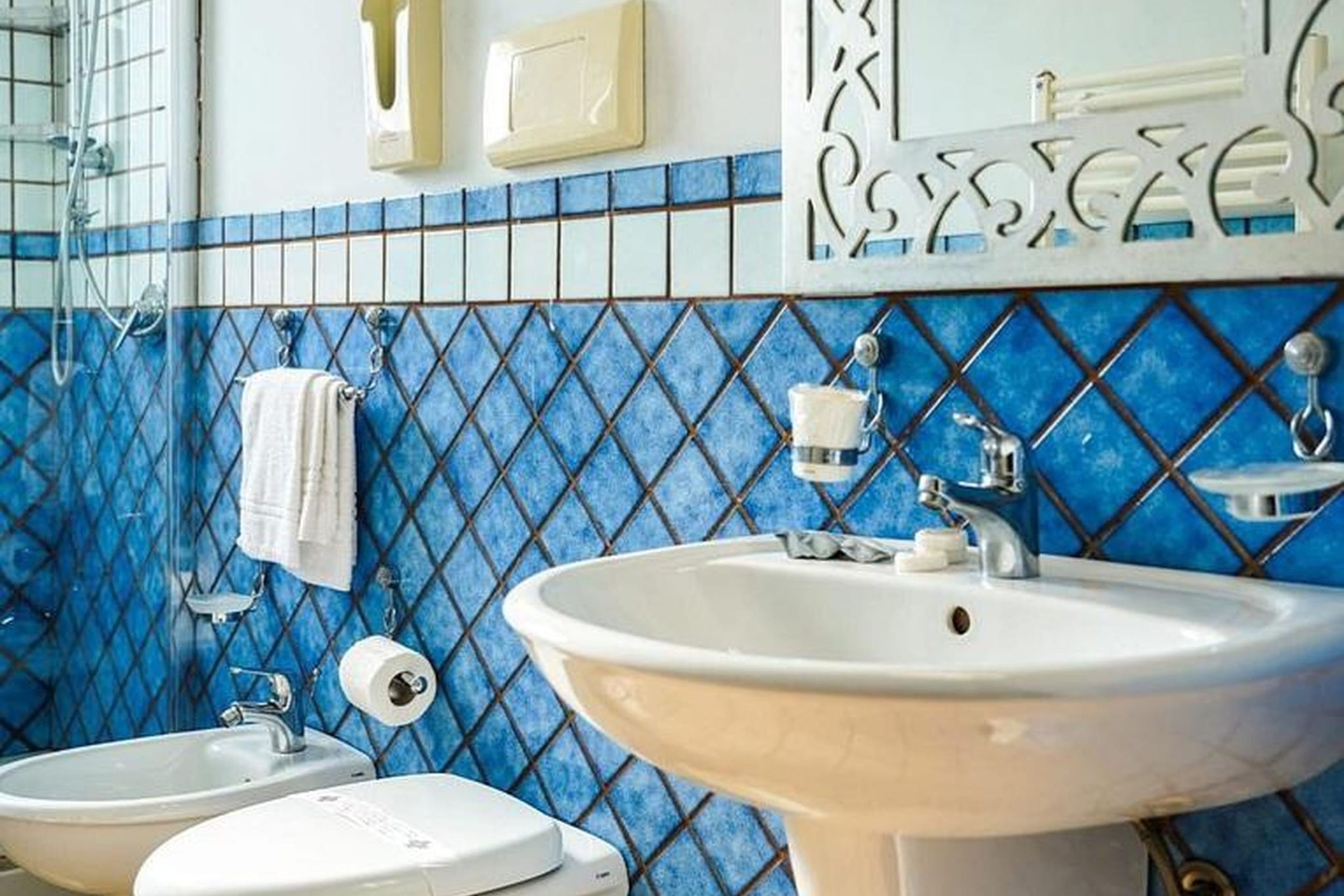 Un baño con un lavabo blanco, dos inodoros, un perchero para toallas y una pared con azulejos azules.