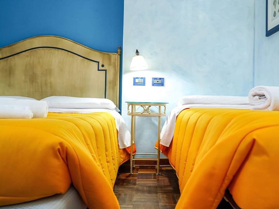 A hotel room with two beds, each with a yellow bedspread and white towels, a wooden headboard, a small table with a lamp, and a blue wall.