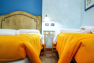 Camera d'albergo con due letti, ognuno con copripiumino giallo e asciugamani bianchi, una testiera in legno, un tavolino con una lampada e una parete blu.