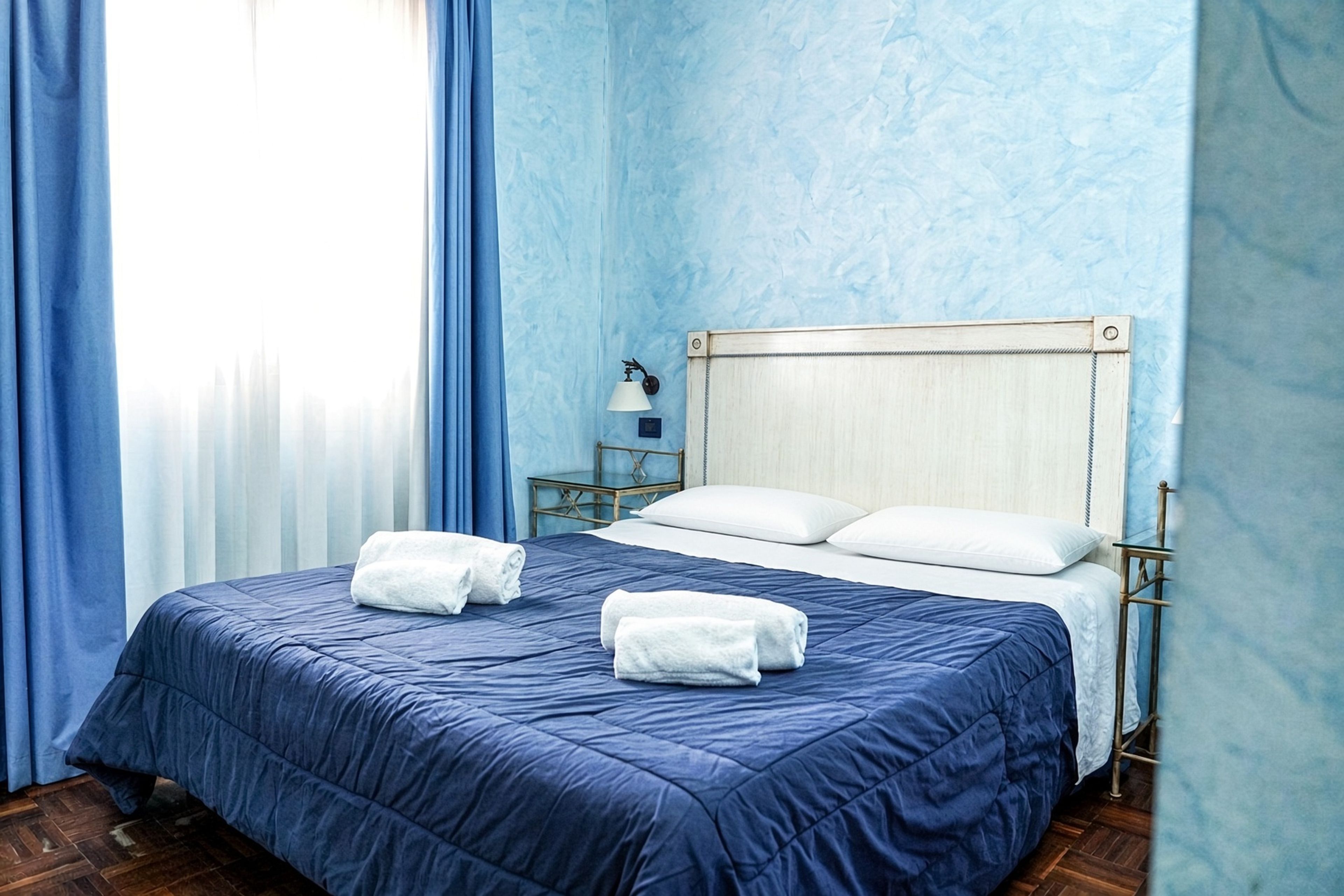 A hotel room with a bed, two pillows, a nightstand, a lamp, and a window with curtains.