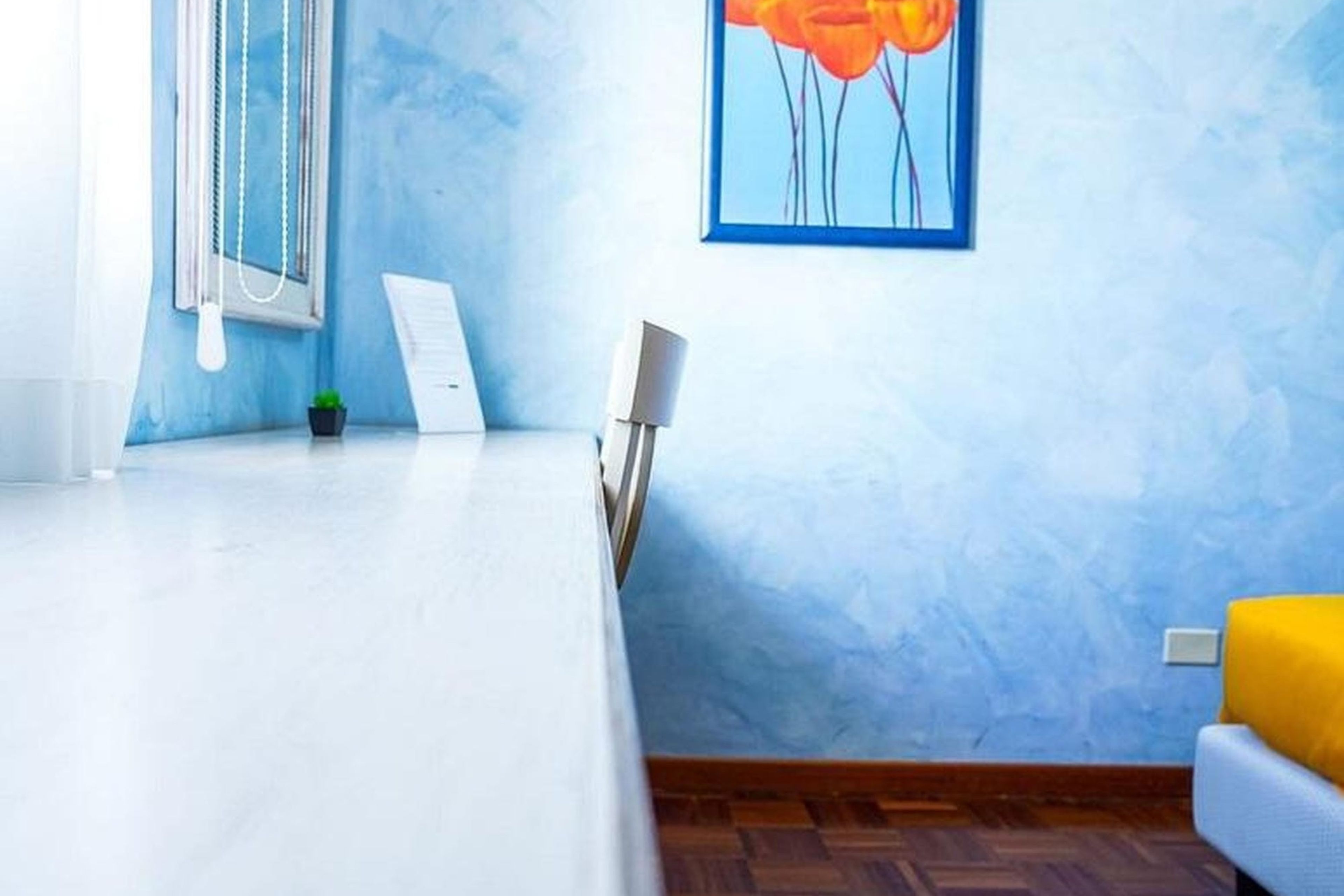 Une pièce avec un lit blanc, une petite table, une chaise, une plante en pot et un tableau accroché au mur.