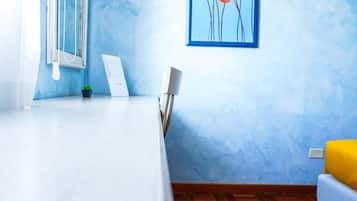 Une pièce avec un lit blanc, une petite table, une chaise, une plante en pot et un tableau accroché au mur.