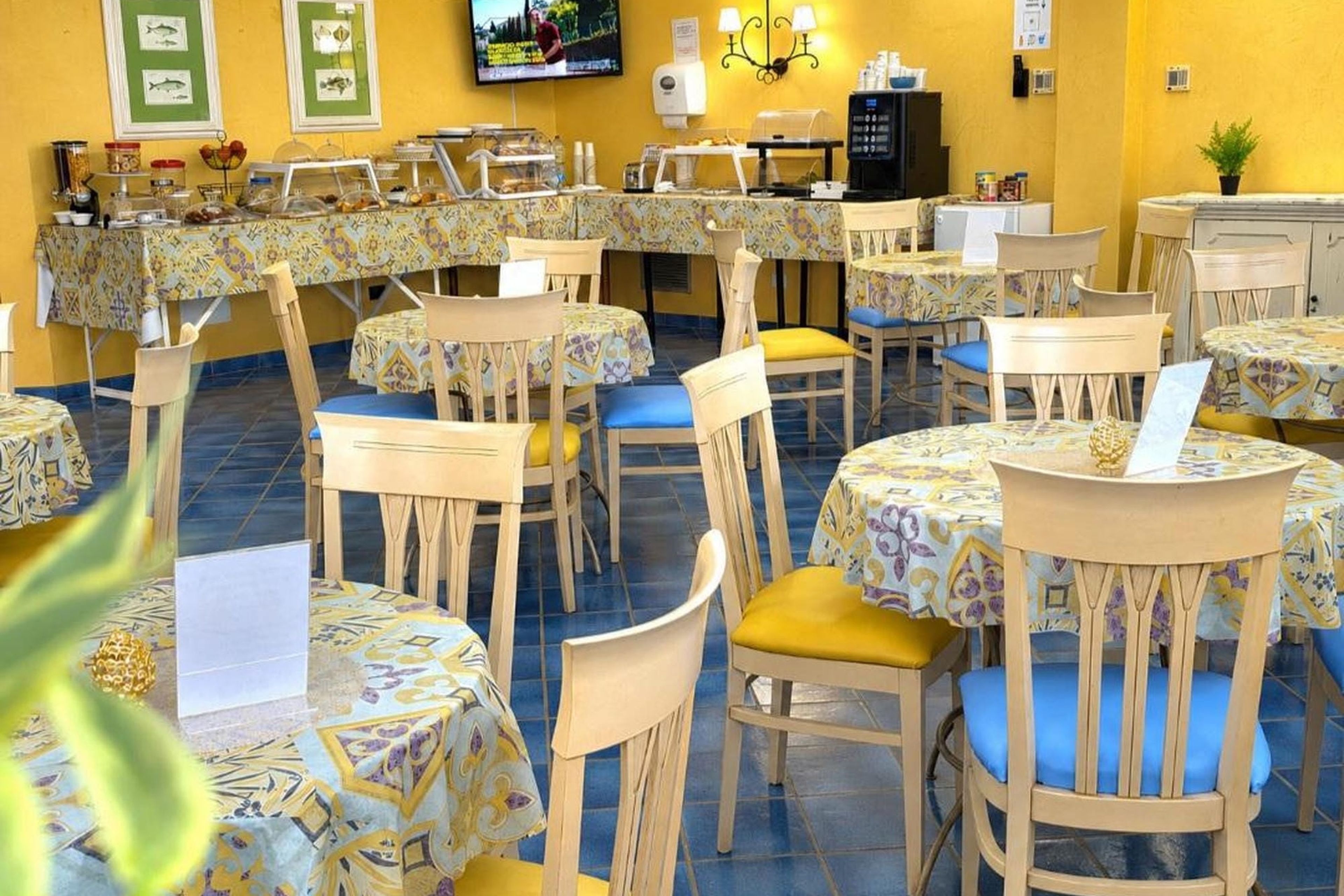 Een restaurant met meerdere tafels en stoelen, een aan de muur bevestigde televisie, en een koffieapparaat.