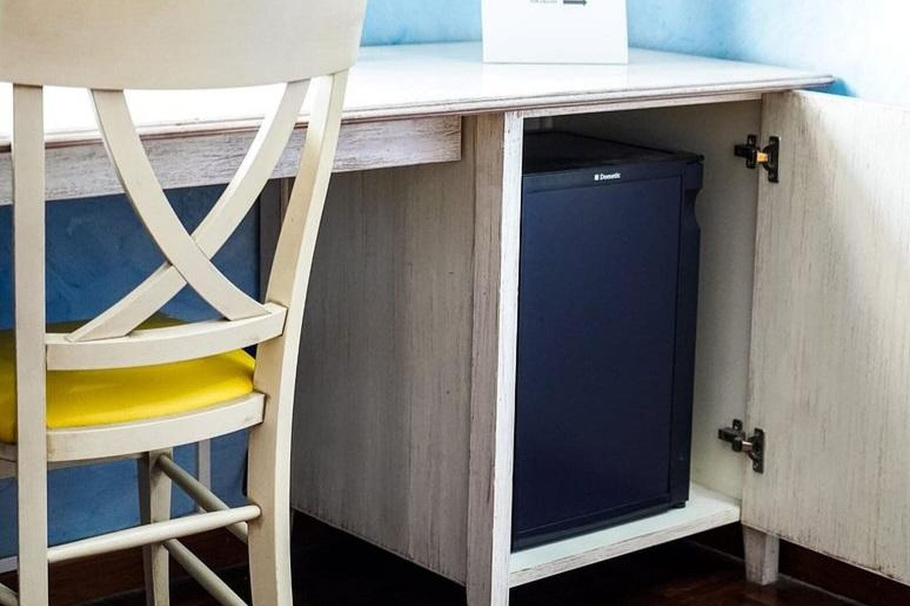 A compact, dark-colored refrigerator in a wooden cabinet with a metal handle, a yellow shelf, and a white chair with a metal frame.