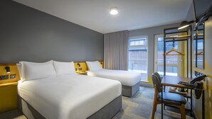 Ein Hotelzimmer mit zwei Betten, einem Schreibtisch, einem Stuhl, einem Kleiderhaken und einem Fenster mit Blick auf die Straße.