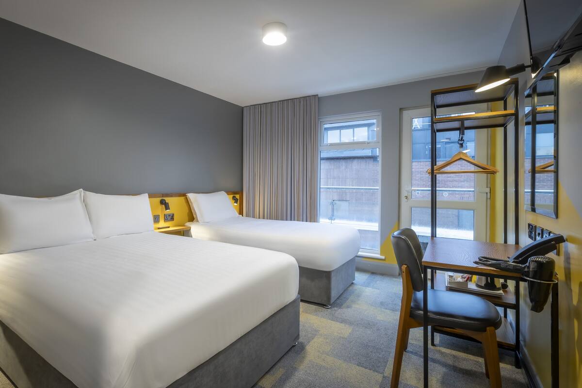 A hotel room with two beds, a desk, a chair, a hanging coat rack, and a window with a view of the street.