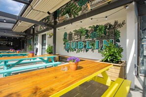 Un espace extérieur aménagé avec des tables et des bancs en bois, une fresque murale représentant le mot « DUBLIN » ainsi que des illustrations de bâtiments et de plantes, et des lampes suspendues.