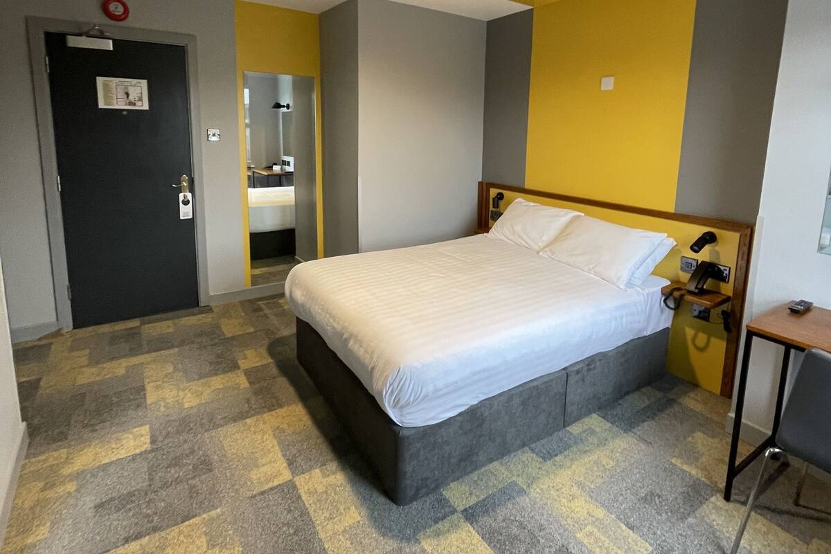 A hotel room with a bed, a desk, a chair, a mirror, and a door.