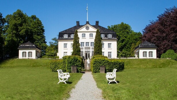 A large, symmetrical building with a central entrance, flanked by two smaller structures, set in a well-manicured lawn with a gravel path leading to the entrance.