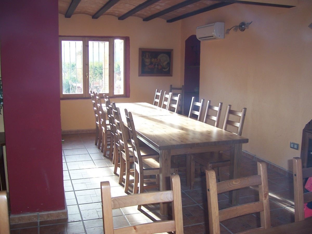 A dining area with a long wooden table and chairs, a red pillar, and a window with a view of greenery.