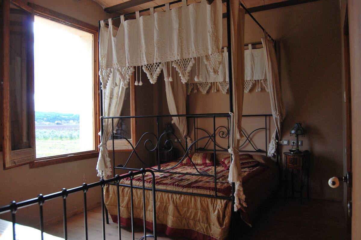 A four-poster bed with a canopy, a bedside table, and a view of the outdoors through a window.
