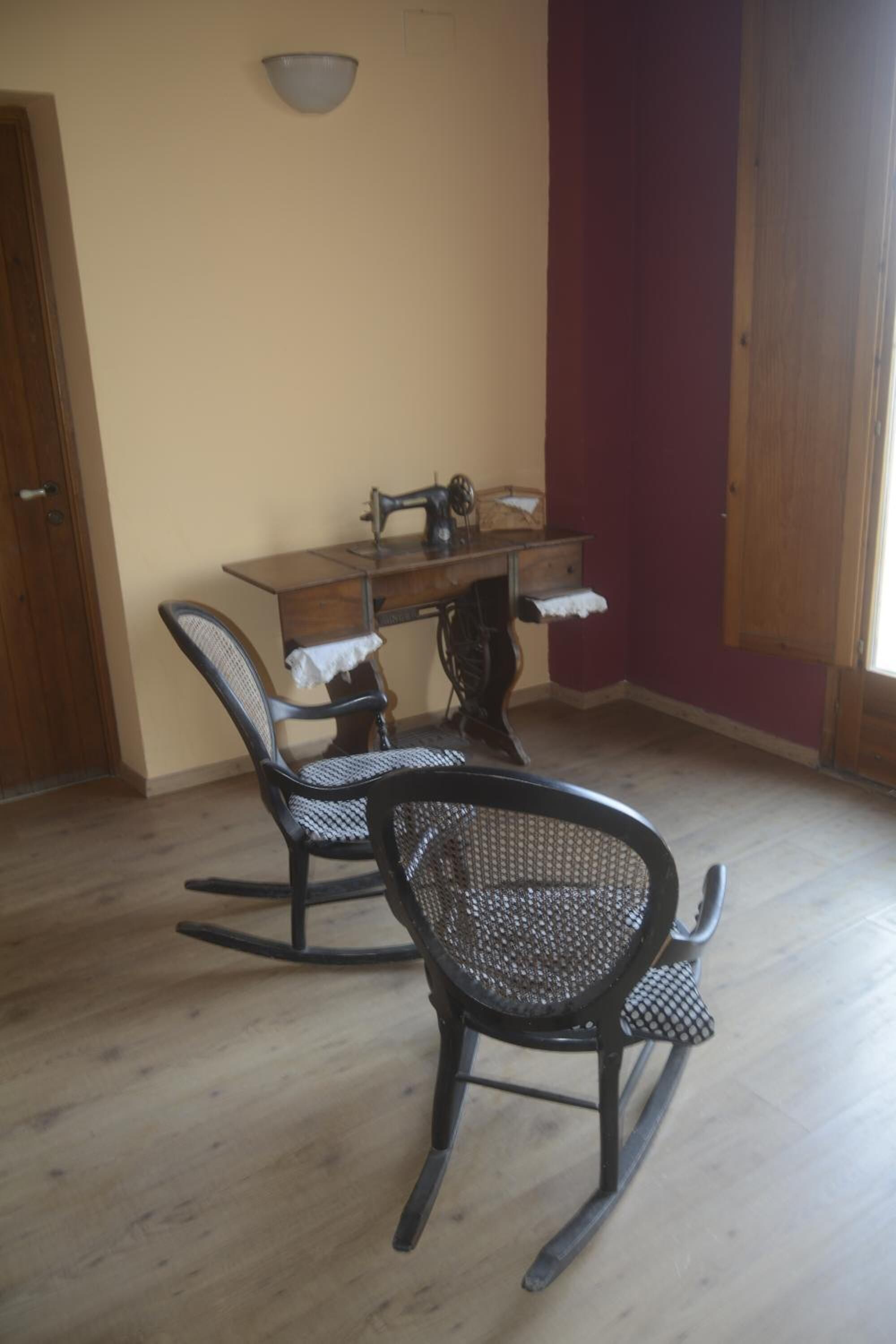A room with a wooden dining table, two rocking chairs, a sewing machine, and a light fixture on the wall.