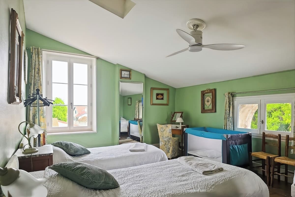 A room with two beds, a ceiling fan, a window with a view, and a chair.